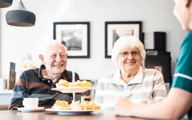 an elderly couple eating cakes in a residential care home with a nurse